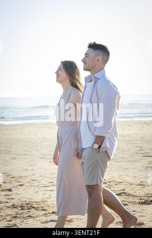 Couple enjoys a walk on the beach during sunny day near ocean waves and sandy shore while holding hands and smiling Stock Photo