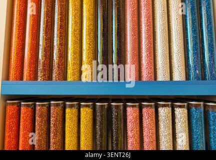Many small chocolate candies with glossy coating are stored in clear jars. These jars are placed on a blue shelf in a candy shop. Various colors fill Stock Photo