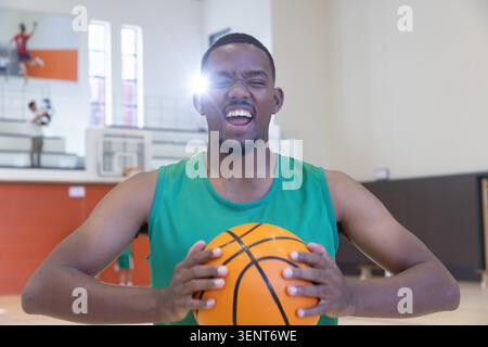 Adult African American man shouting, holding orange basketball in green jersey on hardwood court Stock Photo