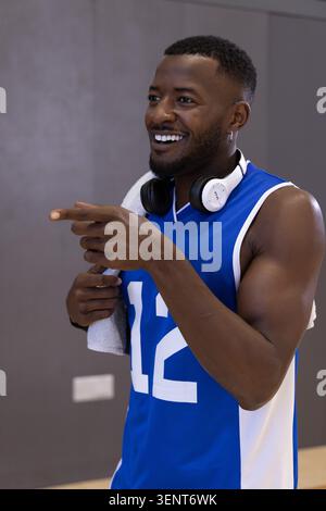 Man in blue jersey #12 holding towel, wearing headphones, smiling, pointing left at gym Stock Photo
