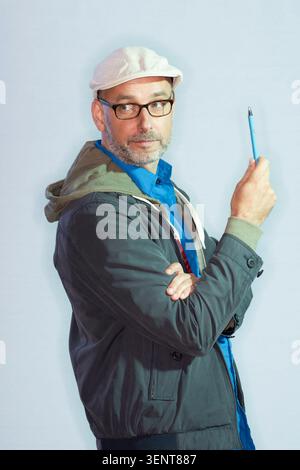 Reinhard Kleist stands in a studio environment, holding a blue pen. He wears glasses and a hat, with a serious look as he engages in creative thinking Stock Photo