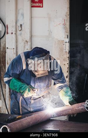 A skilled industrial welder wearing full protective gear, including a mask and heavy-duty gloves, performs precision arc welding on a metal pipe at an Stock Photo