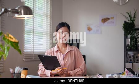 Young Asian creative professional smiling with clipboard in contemporary home office with natural light and plants Stock Photo