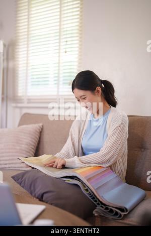 Smiling young Asian woman choosing fabrics and colors for interior design project sitting on couch with material samples Stock Photo