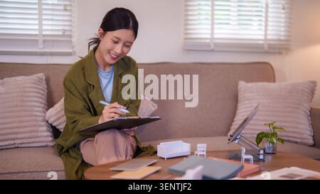 Young Asian female designer drawing furniture sketches and planning project in modern living room with samples Stock Photo