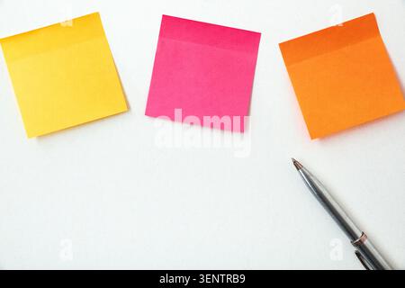 Yellow, pink and orange sticky notes with a silver pen on a white desk. Creative planning, note-taking and office productivity concept. Stock Photo