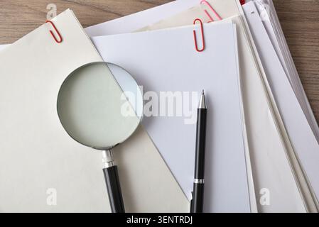Close-up of a magnifying glass resting on stacked documents and blank papers on a desk, symbolizing inspection, review, research, and office organizat Stock Photo