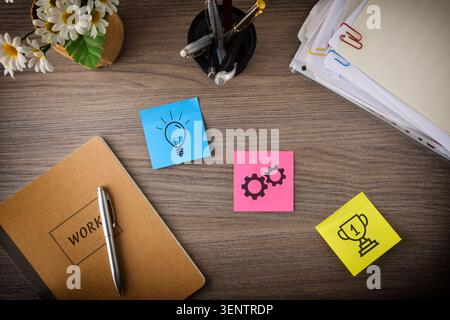 Top view of a wooden desk with notebook, pens, documents, flowers, and colorful sticky notes showing idea, process, and achievement concepts. Stock Photo