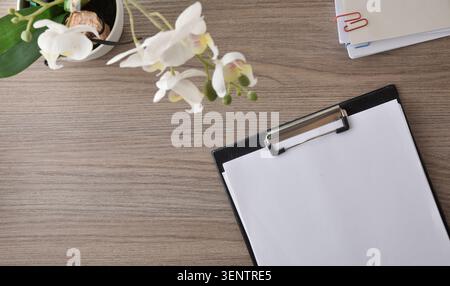 Minimal workspace featuring a blank clipboard, white orchid, notebooks, and paper clips on a wooden desk. Stock Photo