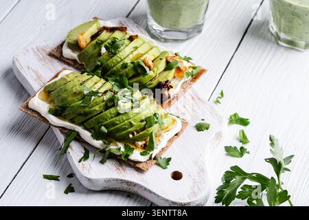 Rye thin crackers with sliced avocado and cream cheese, glass of avocado smoothie on white wood background Stock Photo