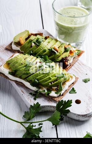 Rye thin crackers with sliced avocado and cream cheese, glass of avocado smoothie on white wood background Stock Photo
