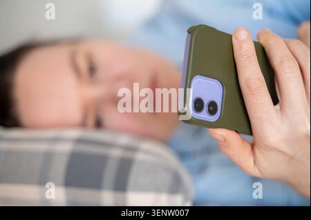 Woman lying on bed in blue shirt after work scrolling through smartphone, home lifestyle and relaxation Stock Photo