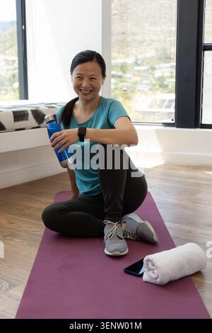 Asian middle-aged woman sitting on mat on hardwood floor smiling in teal shirt holding blue bottle Stock Photo