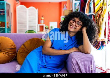 Young hispanic woman with curly hair and eyeglasses sitting comfortably on a vibrant sofa, smiling and enjoying a relaxed moment in her colorful and b Stock Photo