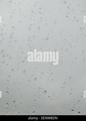 Raindrops on the surface of window panes with gray gloomy cloudy background. Black and white vertical photo. Natural pattern of water droplets. Stock Photo