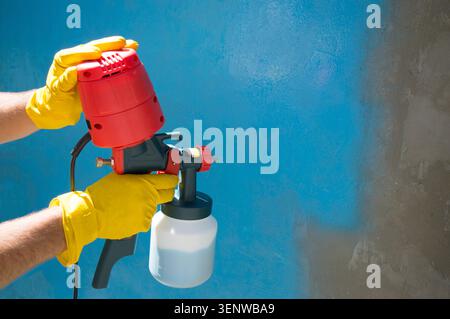 A worker's hands in yellow gloves holds a hand-held spray gun with an overhead compressor. Painting the wall of the house in blue. Household Stock Photo