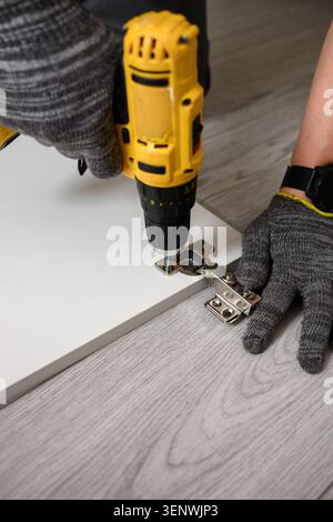 Installing concealed metal hinge with electric screwdriver furniture assembly process close up worker in gloves using cordless power drill carpentry r Stock Photo