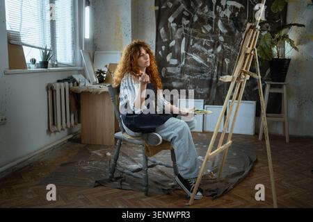 Thoughtful curly haired woman artist thinking about idea for new painting in creative studio. Creative block, inspiration search, imagination and arti Stock Photo