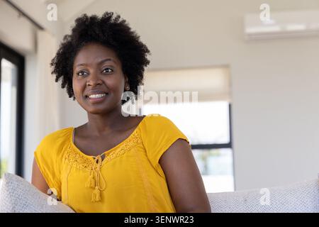 African American woman sitting on light sofa by window with cushion, in yellow tassel blouse Stock Photo