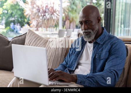 Mature African man working on silver laptop, sitting on beige sofa near glass doors, copy space Stock Photo