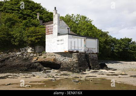A wider perspective of the historic white bathing house sitting above the rocky shoreline and sandy beach at Penrhos Coastal Park, Wales. Stock Photo