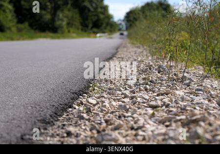 Asphalt road surface meeting a gravel shoulder and green wild plants along a country roadside, with blurred trees and an approaching car in the distan Stock Photo