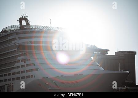Telephoto close view of a cruise ship bow and upper decks backlit by intense sun, with circular lens flare, bright haze and port building silhouette Stock Photo