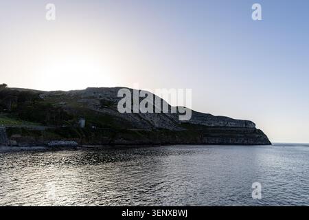 A serene coastal scene featuring a dramatic cliffside at sunset, with calm waters reflecting the sky at Great Orme, in Llandudno, North Wales. Stock Photo