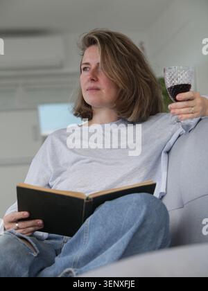 Relaxed woman reading a book while enjoying a glass of wine on a sofa at home. Cozy lifestyle, leisure time, self care and quiet evening concept. Stock Photo