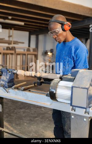 Woodworker shaping wood spindle on lathe in workshop with chisels, wearing goggles and headset Stock Photo