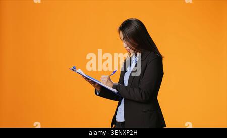 Manager writes important data on papers with clipboard, taking notes to create full report in front of orange background, with young woman in business clothing filling in documents, keeping track. Stock Photo
