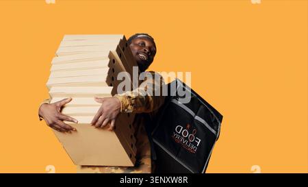 Young deliveryman giving huge stack of pizza boxes to client, delivering big fast food order and carrying takeaway backpack, with male model posing as delivery person with takeout food. Stock Photo