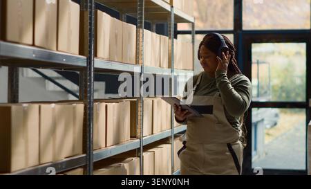 Joyful warehouse chief supply chain officer listening music while inspecting merchandise, cheerful African American logistics coordinator wearing headphones while walking through repository rows. Stock Photo