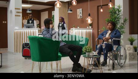 African American guest talking to man while she sits in wheelchair, entering disability friendly hotel lobby, couple travelling abroad, reduced mobility tourism industry. Stock Photo