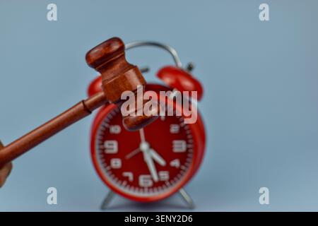 Red alarm clock sits on soft blue surface. Judge hammer rests beside it, highlighting time. Hands point to a late hour, suggesting urgency. Minimal ba Stock Photo