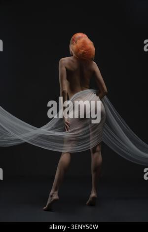 A vibrant red-haired woman poses artistically without clothing, draped in delicate fabric Stock Photo