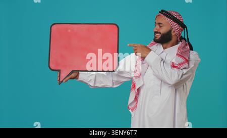 Muslim man shows speech bubble on camera, creating advertisement for new web commercial, young guy working in marketing industry with isolated cardboard sign, empty billboard. Stock Photo