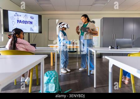 African American teacher holding tablet guiding school-age boy wearing VR headset in classroom Stock Photo