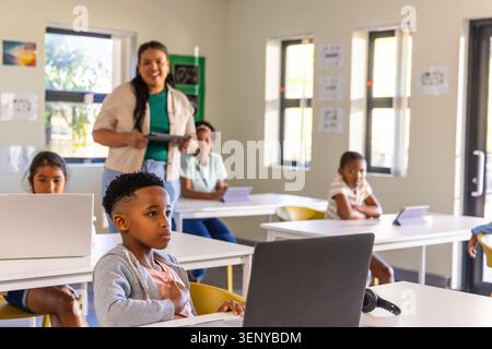 Female teacher holding tablet instructing students in class while African American boy using laptop Stock Photo