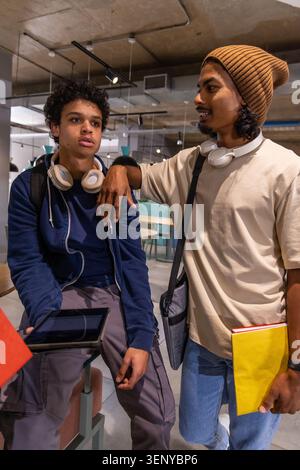 Diverse male students sitting in campus lounge wearing hoodie and headphones, sharing tablet Stock Photo