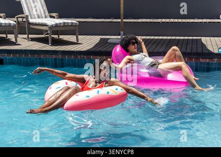 African American women floating on donut and ring floats in home pool in swimsuits and sunglasses Stock Photo