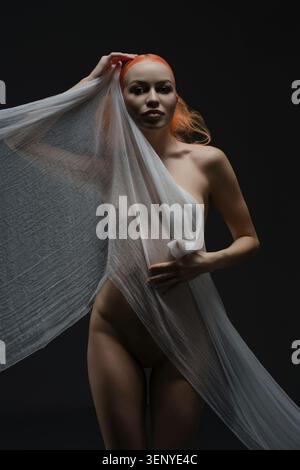 A confident red-haired woman in sheer fabric poses against a dark backdrop, exuding beauty and elegance Stock Photo