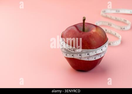 A red apple wrapped in a white measuring tape on a pink background. Weight loss, dieting, nutrition, and healthy lifestyle goals concept. Copy space Stock Photo