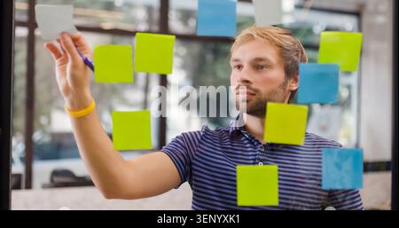 A man works on a whiteboard covered with colorful sticky notes in an office. He writes with a pen while thinking about project tasks and plans. It is Stock Photo