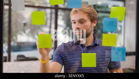 A man stands near a glass wall covered with colorful sticky notes. He focuses as he writes on a note, planning out ideas in a busy office setting. The Stock Photo