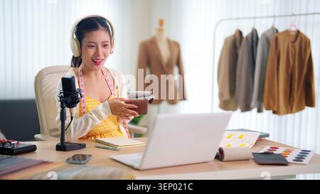 Asian woman hosting fashion design podcast using tablet and microphone at home studio desk with laptop and garment Stock Photo