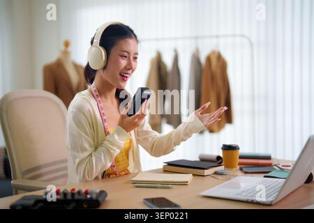 Asian woman podcaster recording audio content on phone in creative home studio with fashion garments background Stock Photo