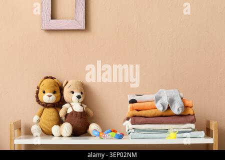 Crochet lion and bear stuffed animals with stacked baby clothes and toys on white shelf against peach wall, the background is a plain peach-colored wa Stock Photo