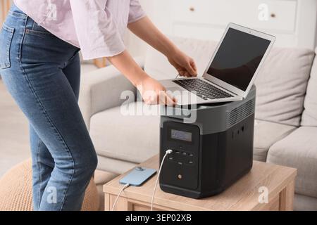 Woman connecting laptop to portable power station for charging, sustainable energy solution for home and travel, the power station also charges a smar Stock Photo