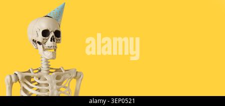 Human skeleton model wearing blue polka dot party hat on bright yellow background with copy space, the skeleton is positioned on a bright yellow backg Stock Photo
